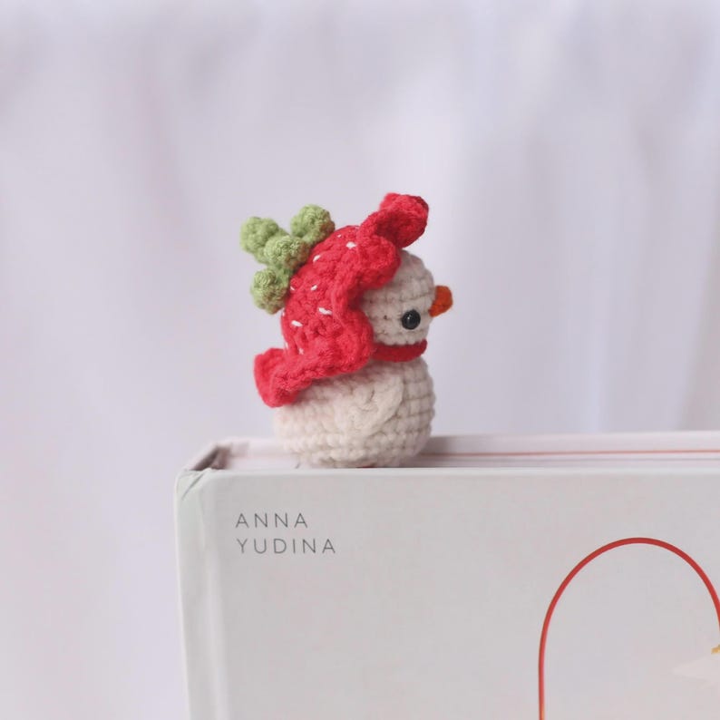 May include: A crocheted snowman figurine with a red hat and green accents. The snowman has a small orange nose and black eyes. The figurine is sitting on a white surface with the text "ANNA YUDINA" visible.