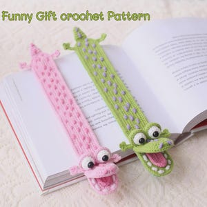 以下が含まれることがあります： ピンクと緑の手編みワニのブックマークが2つ、開いた本の上に置かれています。ピンクのワニは口を開けており、緑のワニは口を閉じています。「Funny Gift crochet Pattern」の文字が上部にあります。