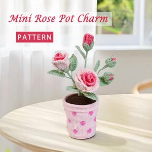 Könnte beinhalten: Gehäkelter Mini-Rosen-Topf-Charm mit rosa Rosen und grünen Blättern. Der Topf ist hellrosa mit einem Tupfenmuster. Der Text "Mini Rose Pot Charm" und "PATTERN" ist im Bild zu sehen.