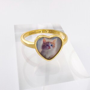Op de afbeelding: Een goudkleurige ring met een hartvormig gezicht met een foto van een kat. De ring heeft de inscriptie "Lovely cat Ella 1-2025" gegraveerd op de band. De ring wordt gepresenteerd op een helder, rechthoekig blok.