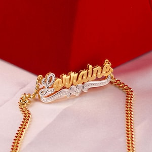 以下が含まれることがあります： 「Lorraine」という名前が重ねてデザインされたゴールドカラーのネックレス。名前はゴールド仕上げの筆記体フォントで、シルバーのベースにセットされています。ネックレスにはハートのディテールとゴールドチェーンが付いています。