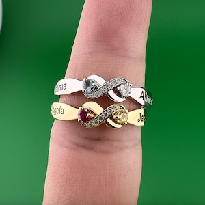 Puede incluir: Dos anillos con forma de infinito, uno plateado y otro dorado, cada uno con una piedra preciosa y nombres grabados. El anillo plateado tiene una piedra azul y otra transparente, mientras que el dorado tiene una roja y una amarilla.