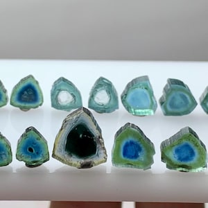 Op de afbeelding: Een verzameling watermeloen toermalijn plakjes, met een levendige mix van kleuren. De edelstenen tonen een groene buitenlaag, een witte of heldere middenlaag en een blauw centrum. Deze gepolijste stenen zijn gerangschikt op een wit oppervlak.