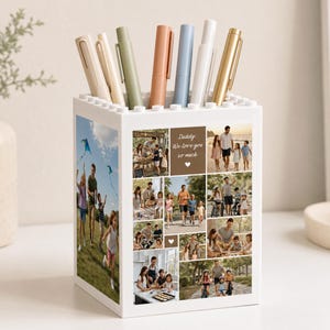 Puede incluir: Un portalápices cuadrado blanco con un borde superior de bloques de construcción, que presenta un collage de fotos familiares en los paneles frontal y lateral. El texto "Daddy, We love you so much" está impreso en el frente. Varios bolígrafos están guardados.
