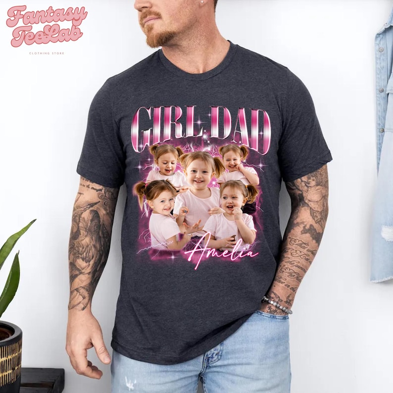 K&ouml;nnte beinhalten: Dunkelgraues T-Shirt mit dem Text "GIRL DAD" in einem rosa-wei&szlig;en Design. Darunter befindet sich ein Foto von l&auml;chelnden M&auml;dchen mit dem Namen "Amelia" in rosa Schrift. Das Shirt stammt vom Bekleidungsgesch&auml;ft "Fantasy TeeLab".