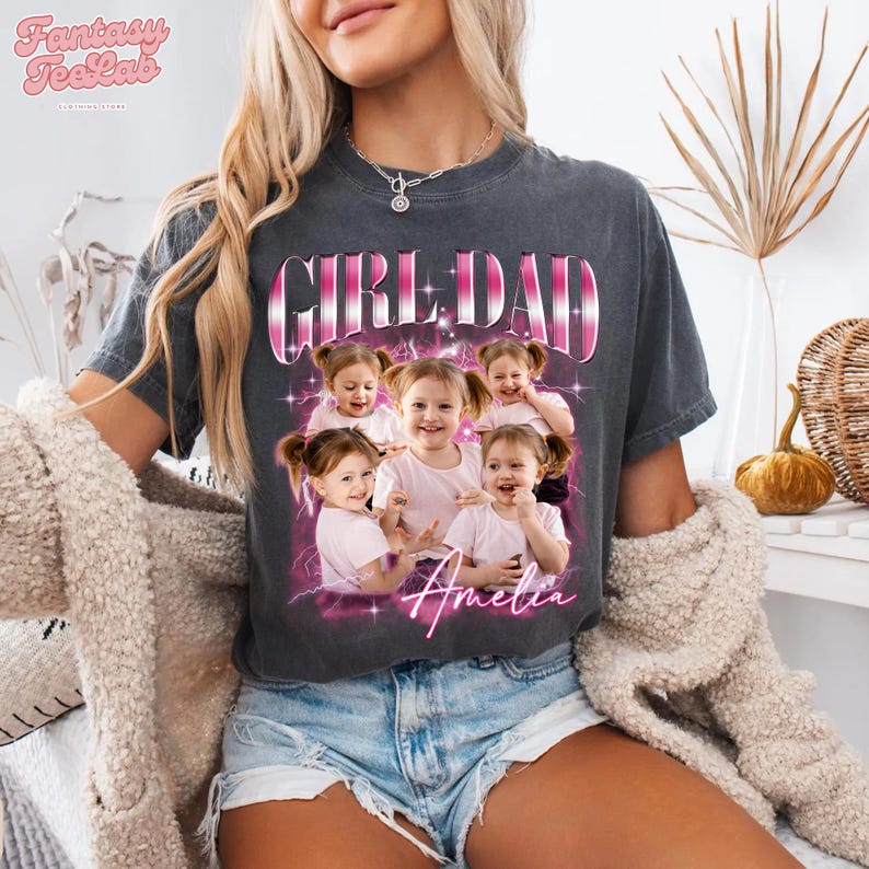K&ouml;nnte beinhalten: Dunkelgraues T-Shirt mit dem Schriftzug "GIRL DAD" in Rosa und Wei&szlig;, mit einem Foto von sechs jungen M&auml;dchen. Der Name "Amelia" steht in rosa Schrift darunter. Das Shirt wird mit Jeansshorts getragen.