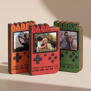 May include: Three wooden photo frames shaped like retro game consoles. The frames are orange, red, and green, each with a different photo and the words "DADDY", "PAPA", and "DADDY" across the top. Each frame has the text "THANKS FOR ALWAYS LEVELING US UP".