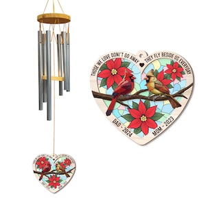 May include: A wind chime with silver tubes and a wooden top, alongside a heart-shaped ornament. The ornament features two birds, red poinsettias, and the text "THOSE WE LOVE DON'T GO AWAY THEY FLY BESIDE US EVERYDAY" with "DAD-2024" and "MOM-2023".