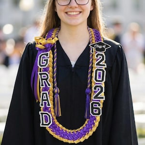 Puede incluir: Un lei de graduación con las palabras "GRAD" y "2026" en letras blancas con bordes negros. El lei es morado y dorado, con borlas y cintas a juego. La persona lleva una toga de graduación negra.