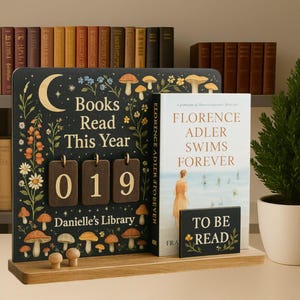 Puede incluir: Un elemento decorativo de estantería de madera con un fondo oscuro, que presenta el texto "Books Read This Year" y el número "019". El diseño incluye ilustraciones de setas y flores. Un libro titulado "Florence Adler Swims Forever" se muestra junto a él, junto con un letrero de "To Be Read".