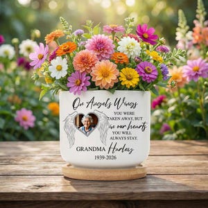 Puede incluir: Una maceta de cerámica blanca con flores coloridas. La maceta tiene una foto en forma de corazón y el texto "On Angel's Wings" y "Grandma Harley 1939-2026". Se encuentra sobre un posavasos de madera.