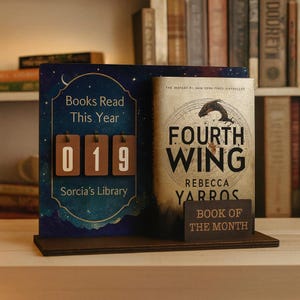 May include: A wooden book display with a book and a counter. The counter reads "019" for books read this year. The book is titled "Fourth Wing" by Rebecca Yarros. A smaller sign reads "Book of the Month". The background is a bookshelf.