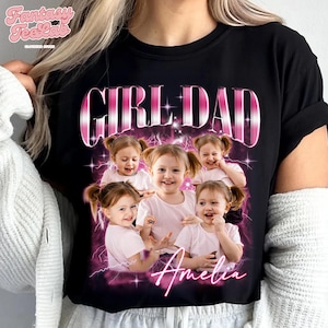 K&ouml;nnte beinhalten: Schwarzes T-Shirt mit dem Text "GIRL DAD" in Rosa und Wei&szlig;, mit einer Fotocollage eines jungen M&auml;dchens. Der Name des M&auml;dchens, Amelia, ist darunter in Kursivschrift geschrieben. Das Shirt hat einen Rundhalsausschnitt.