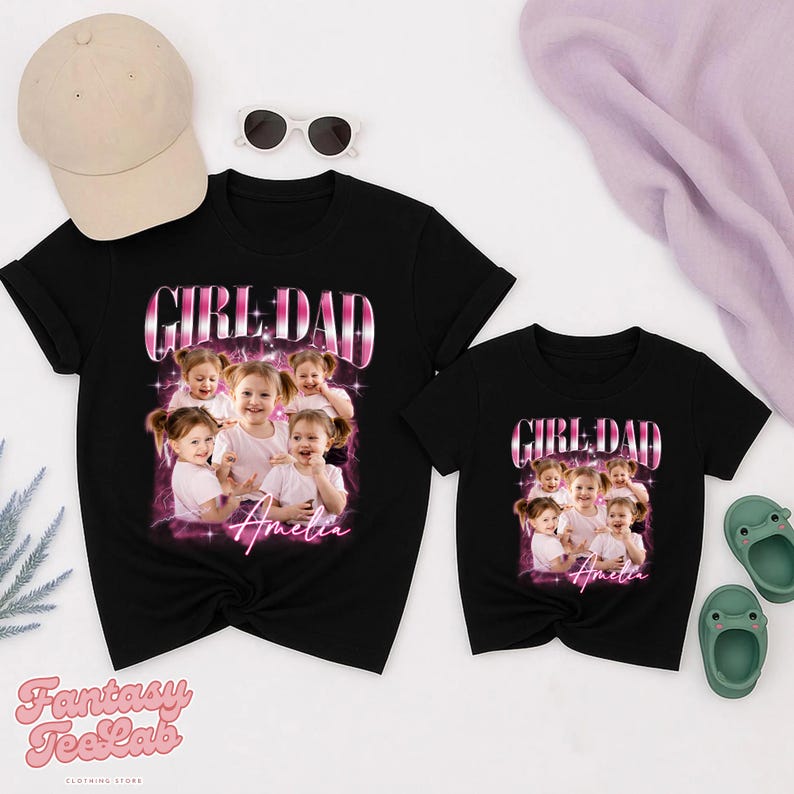 K&ouml;nnte beinhalten: Zwei schwarze T-Shirts mit dem Text "GIRL DAD" in einem rosa-wei&szlig;en Design, mit einem Foto eines jungen M&auml;dchens. Der Name "Amelia" ist unter dem Foto geschrieben. Eine beige Baseballkappe, Sonnenbrille und gr&uuml;ne Sandalen sind ebenfalls im Bild.