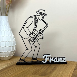 Könnte beinhalten: Schwarz-weiße Linienkunst-Skulptur eines Saxophonspielers in Anzug und Hut. Die Skulptur steht auf einem schwarzen Sockel mit dem Namen "Franz" in weißer Schrift. Im Hintergrund eine weiße Vase mit dekorativen Blumen.