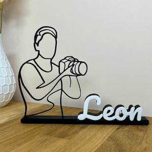Può includere: Scultura in linea bianca e nera di una persona che tiene una macchina fotografica. Il nome "Leon" è visualizzato in lettere bianche in corsivo su una base nera. La scultura è su una superficie di legno.
