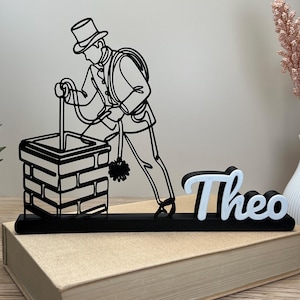 Könnte beinhalten: Eine Schwarz-Weiß-Linienkunst-Skulptur eines Schornsteinfegers mit dem Namen "Theo" in Weiß und Schwarz. Der Schornsteinfeger arbeitet an einem Ziegelschornstein. Die Skulptur steht auf einem Buch.