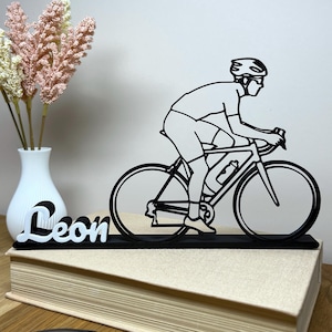 May include: Black metal cyclist silhouette on a bicycle, with the name "Leon" in white script. The cyclist is wearing a helmet and riding gear. The sculpture is displayed on a book, with a white vase and flowers in the background.