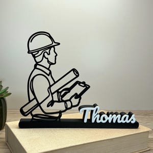 Pode incluir: Silhueta preta e branca de um engenheiro segurando plantas e uma prancheta. O nome "Thomas" é exibido em branco e azul claro. A estatueta é colocada em cima de um livro.
