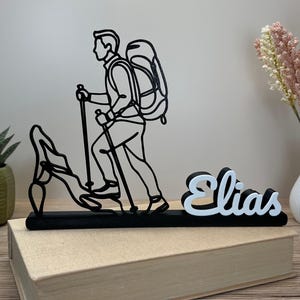 Può includere: Silhouette nera e bianca di un escursionista con zaino e bastoncini da trekking, che scala una montagna. Il nome "Elias" è visualizzato in corsivo bianco e nero. Il pezzo decorativo è posto su un libro chiuso.