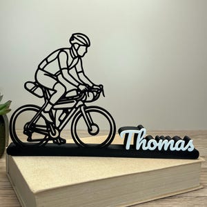 Könnte beinhalten: Schwarz-weißes Schreibtischzubehör zum Thema Fahrrad, das einen Radfahrer auf einem Rennrad zeigt. Der Name "Thomas" wird in Weiß und Hellblau angezeigt. Das Zubehör befindet sich auf einem Buch.