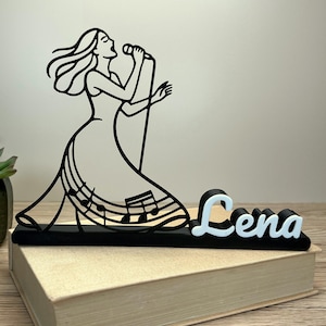 May include: Black and white silhouette of a woman singing into a microphone. The figure is atop a black base with musical notes and the name "Lena" in white and black. The sculpture is placed on a closed book.
