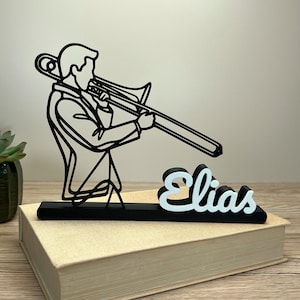 Puede incluir: Silueta en blanco y negro de un trombonista con el nombre "Elias" en fuente cursiva. La escultura está montada sobre una base negra y descansa sobre un libro cerrado. El nombre es blanco con una sombra azul claro.