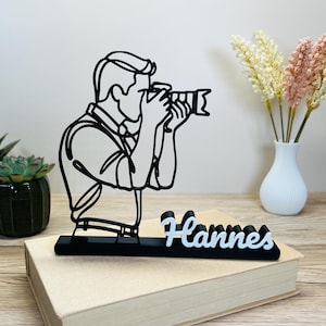 Könnte beinhalten: Schwarz-weiße Linienkunst-Skulptur eines Fotografen mit Kamera. Die Skulptur ist auf einem schwarzen Sockel mit dem Namen "Hannes" in weißer Schrift montiert. Die Skulptur steht auf einem geschlossenen Buch.