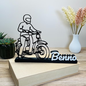 Op de afbeelding: Zwart-wit silhouet van een persoon op een scooter, met de naam "Benno" in het wit. De sculptuur staat op een gesloten boek. Decoratieve vetplanten en bloemen staan op de achtergrond.