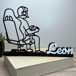 Könnte beinhalten: Schwarz-weiße Gamer-Figur, die eine Person in einem Gaming-Stuhl zeigt, die ein Headset trägt und einen Controller hält. Der Name "Leon" wird in Weiß und Hellblau angezeigt. Die Figur steht auf einem Buch.