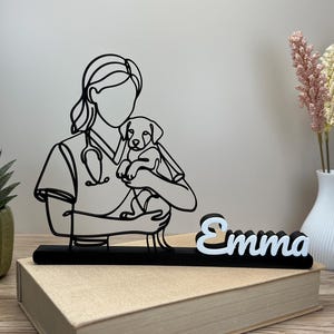 Könnte beinhalten: Eine schwarze Drahtkunstskulptur, die eine Tierärztin mit einem Welpen darstellt, montiert auf einem schwarzen rechteckigen Sockel. Der Name "Emma" ist in weißen 3D-Buchstaben auf der rechten Seite des Sockels angebracht, inklusive Stethoskop.