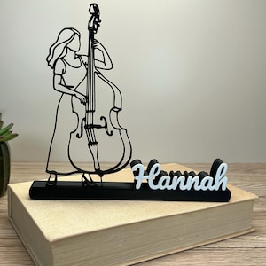 Könnte beinhalten: Eine schwarze Metallskulptur einer Person, die einen Kontrabass spielt. Die Skulptur steht auf einem schwarzen Sockel mit dem Namen "Hannah" in weißer Schrift. Die Skulptur steht auf einem geschlossenen Buch.
