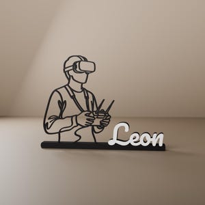 Peut inclure: Une silhouette en art linéaire noir représentant une personne portant un casque de réalité virtuelle et tenant une radiocommande de drone, fixée sur une base noire avec le prénom Leon écrit en lettres cursives blanches sur le côté.