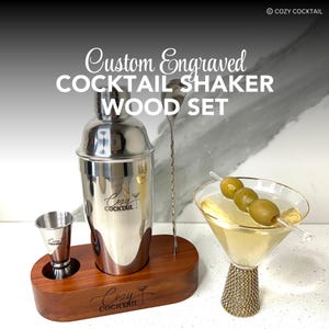 May include: A custom engraved cocktail shaker set with a wooden base. The shaker is stainless steel and has the words "Cozy Cocktail" engraved on it. The set also includes a jigger, a cocktail stirrer, and a martini glass with olives.