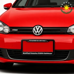 Puede incluir: Un coche Volkswagen rojo con una parrilla negra y un marco de matr&iacute;cula. El marco de la matr&iacute;cula dice "I'D RATHER BE DRIVING A" encima de una matr&iacute;cula roja. Debajo de la placa, dice "Modified Komatsu D355A Bulldozer."