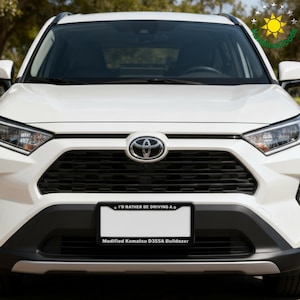 Puede incluir: Un SUV Toyota blanco con una parrilla negra y una placa que dice "I'D RATHER BE DRIVING A" y "Modified Komatsu D355A Bulldozer". El logotipo de Toyota es visible en la parte delantera.