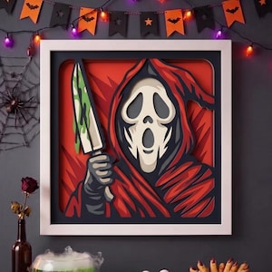 Op de afbeelding: Een vierkant ingelijst meerlaags papierkunstwerk met een figuur in een kap met een wit spookmasker die een groot mes vasthoudt met groene vloeistof die van het lemmet druipt, tegen een levendige rode achtergrond met donkerblauwe en zwarte accenten.