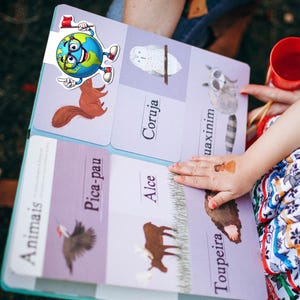 Pu&ograve; includere: Un libro educativo per bambini intitolato Animais con illustrazioni di uno scoiattolo, un gufo, un procione, un picchio, un alce e una talpa. Un personaggio a forma di globo terrestre con occhiali e una bandierina rossa &egrave; fissato in alto.