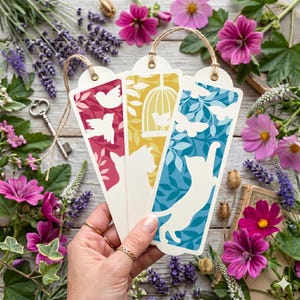 May include: Three decorative bookmarks with bird, cat, and butterfly designs. The bookmarks are cream-colored with red, yellow, and blue backgrounds. Each bookmark has a brown string attached at the top. The image is surrounded by flowers and greenery.