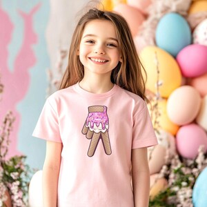 May include: A young girl wearing a pink t-shirt with a graphic of a pink and white ice cream cone on a waffle cone. The girl is smiling and standing in front of a background of pastel colored Easter eggs.