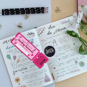 May include: A pink ticket with the text "LIFETIME MEMBER OF THE Cowboy Romance BOOK CLUB" rests on an open journal. The journal pages are filled with handwritten notes, drawings, and stickers. Pencils and paper clips are also visible.