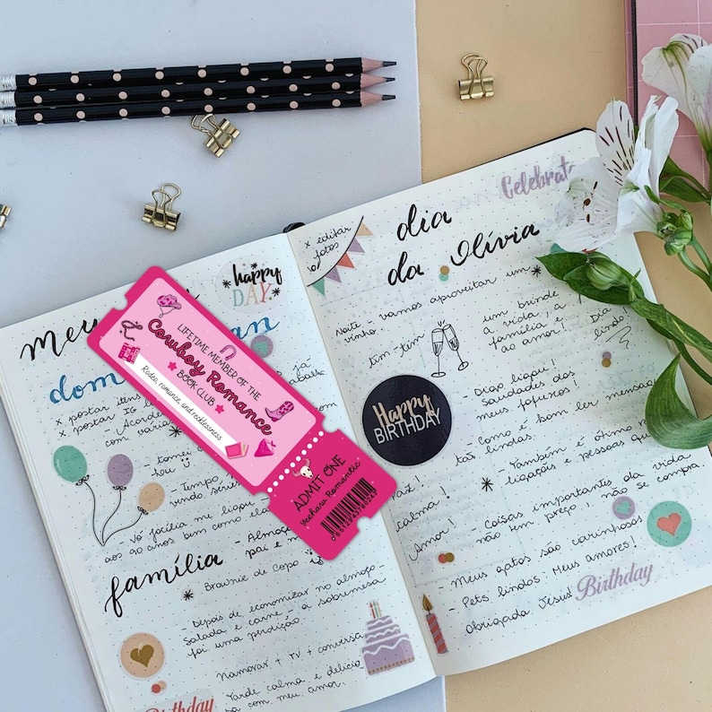 May include: A pink ticket with the text "LIFETIME MEMBER OF THE Cowboy Romance BOOK CLUB" rests on an open journal. The journal pages are filled with handwritten notes, drawings, and stickers. Pencils and paper clips are also visible.