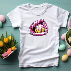 May include: White t-shirt with a pink and yellow softball graphic. The softball is covered in pink frosting and sprinkles.
