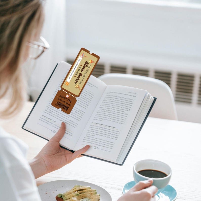 May include: A bookmark shaped like a ticket is placed inside an open book. The bookmark is brown with the text "New Burn Book Club" and "Admit One". A cup of coffee and a plate of food are on the table.