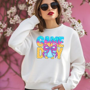 May include: White sweatshirt with a graphic of the words "GAME DAY" in blue, pink, and yellow with a baseball and bow in the center.