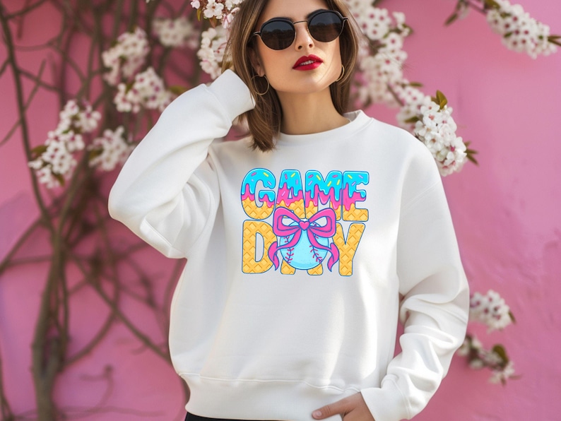 May include: White sweatshirt with a graphic of the words "GAME DAY" in blue, pink, and yellow with a baseball and bow in the center.