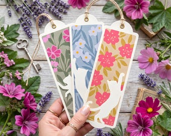May include: Three floral cat bookmarks with beige, blue, and green backgrounds. Each bookmark features a white cat silhouette and a different floral pattern in pink, white, and yellow. The bookmarks have a string loop at the top.