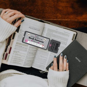 May include: An open book with a bookmark that reads "LIFETIME MEMBER OF THE Dark Romance BOOK CLUB". A small notebook labeled "NOTES" is next to the book. The bookmark has a skull and a ticket that says "ADMIT ONE Danger Lover".