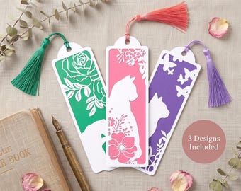 May include: Three rectangular bookmarks featuring intricate laser-cut designs of cats and floral patterns. Each bookmark has a colored background in green, pink, or purple, and is topped with a matching silk tassel. Text on image: 3 Designs Included.