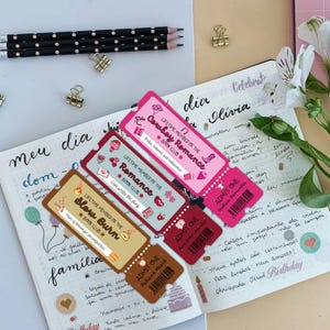 May include: A collection of colorful book club membership cards and admission tickets are displayed on an open journal. The cards feature text like "Cowboy Romance" and "Slow Burn." Pencils and paper clips are also visible.